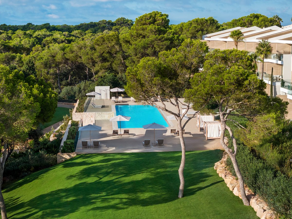 A pool set in a lawn among pine trees, with sun loungers, shaded seating and low resort buildings behind.