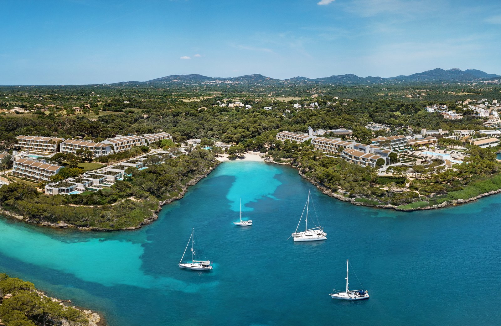 Ikos Porto Petro from the air: low-rise blocks wrapped around a turquoise cove with sailboats moored in it.