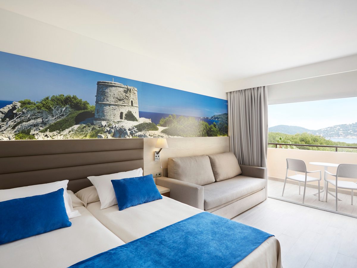 Two single beds and a sofa bed below a painted watchtower mural, with sliding doors to a furnished balcony.