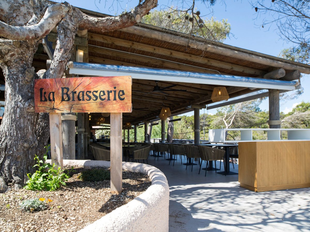 The timber entrance and covered terrace of La Brasserie, its painted wooden sign beside a pine tree.