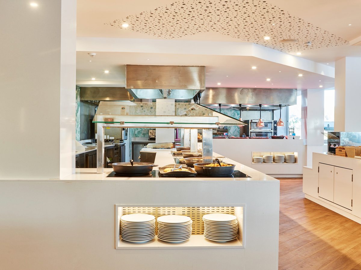 An open kitchen with a stainless-steel extractor hood and stacked plates at a serving counter.