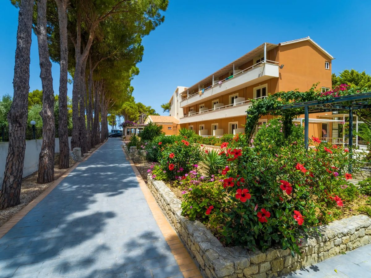 A paved path through the grounds, lined with red-flowering shrubs and tall pine trees, with an accommodation building beyond.