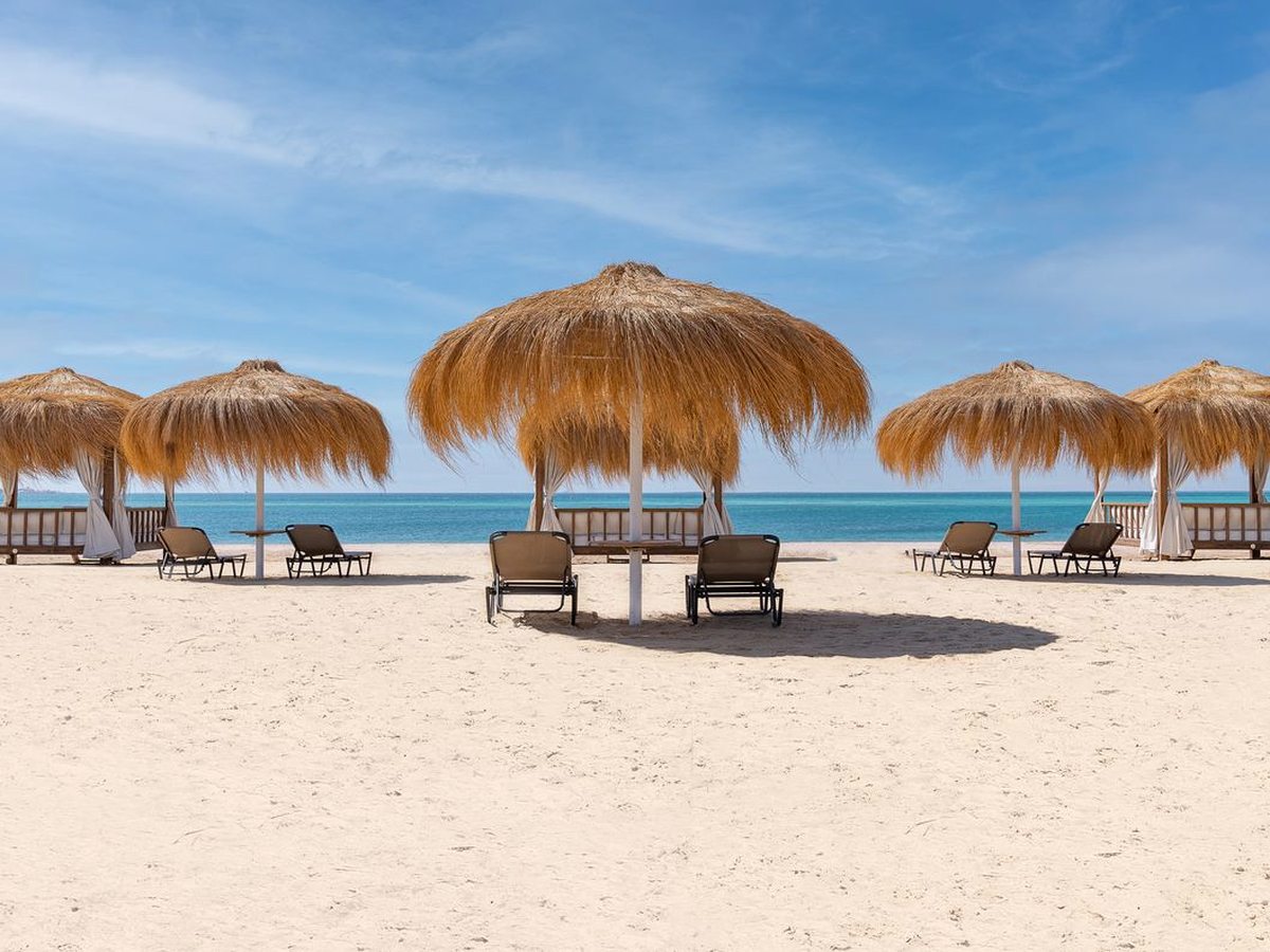 Thatched umbrellas with white-curtained cabanas and dark sun loungers on white sand at the edge of the flat turquoise Red Sea under a blue sky.