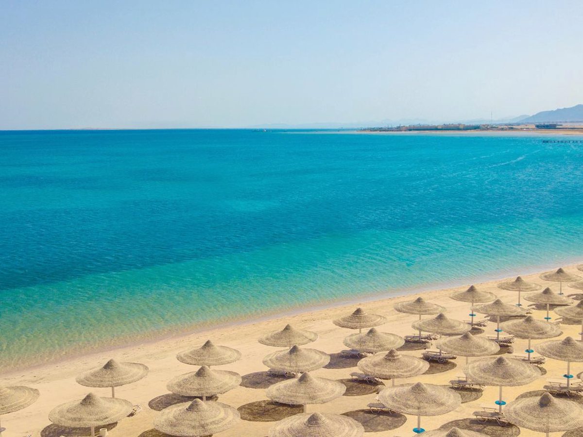 Rows of thatched sun umbrellas on a wide sandy beach beside the calm turquoise Red Sea, with a low desert headland across the bay.