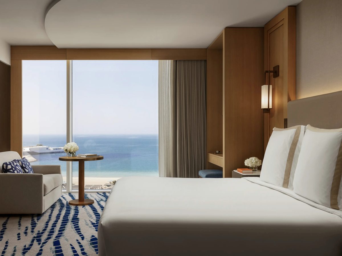 A guest room with a king bed, pale wood panelling and a floor-to-ceiling window looking out over the Arabian Gulf, an armchair and small round table beside it.