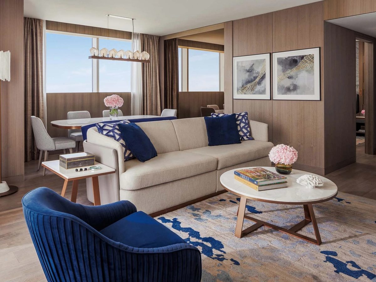 A suite living room with a long cream sofa, a blue velvet armchair, a round marble coffee table and a dining area by tall windows.