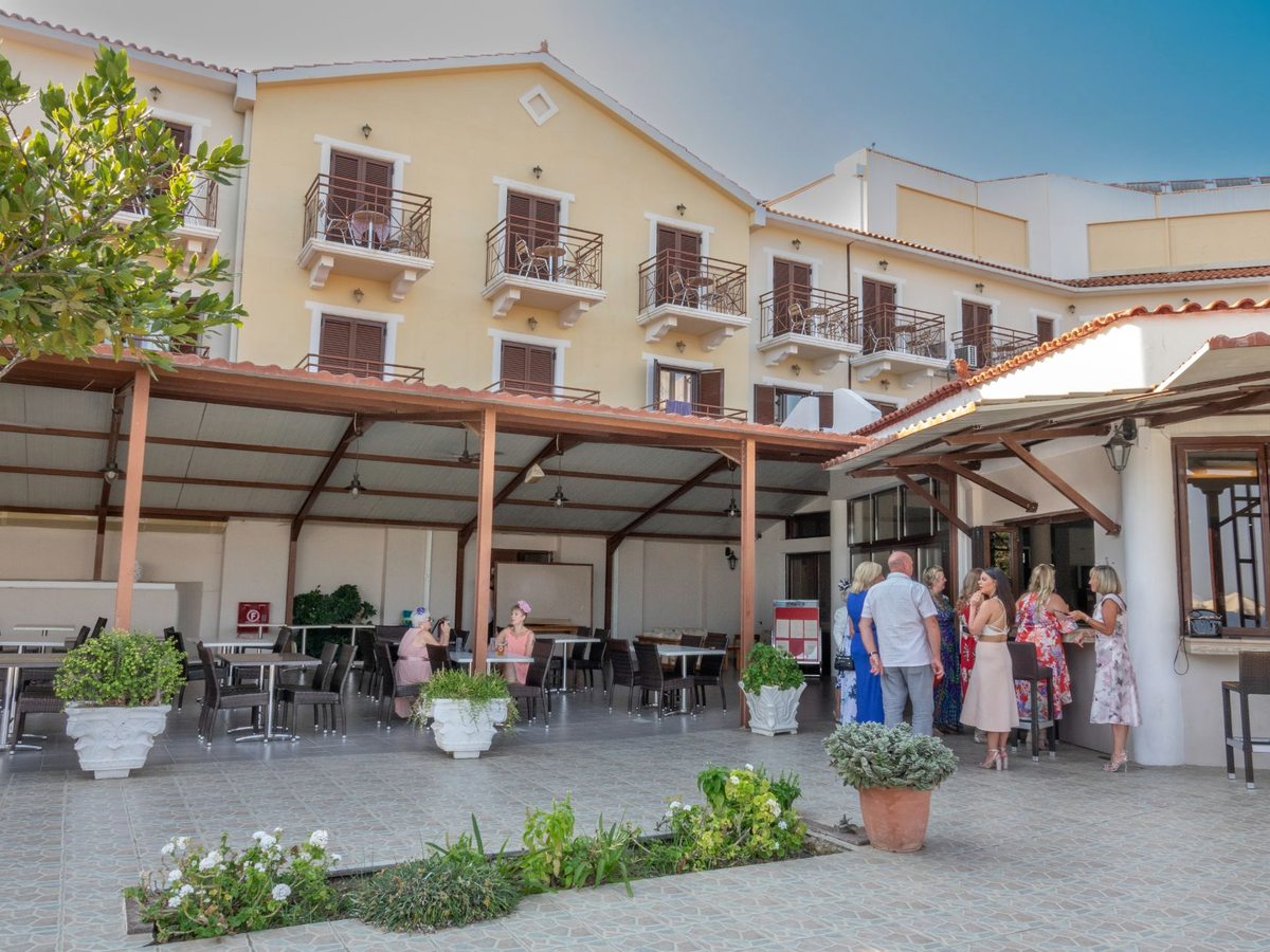 A covered outdoor dining terrace with tables and chairs beside the hotel building, guests standing near the entrance.