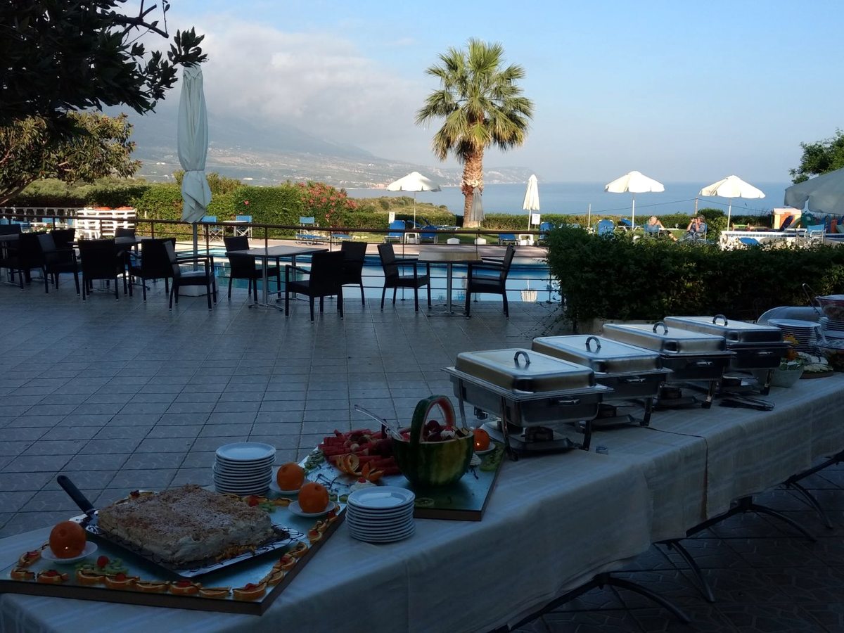 A buffet with metal chafing dishes and stacked plates set on tables on an outdoor terrace overlooking the sea.