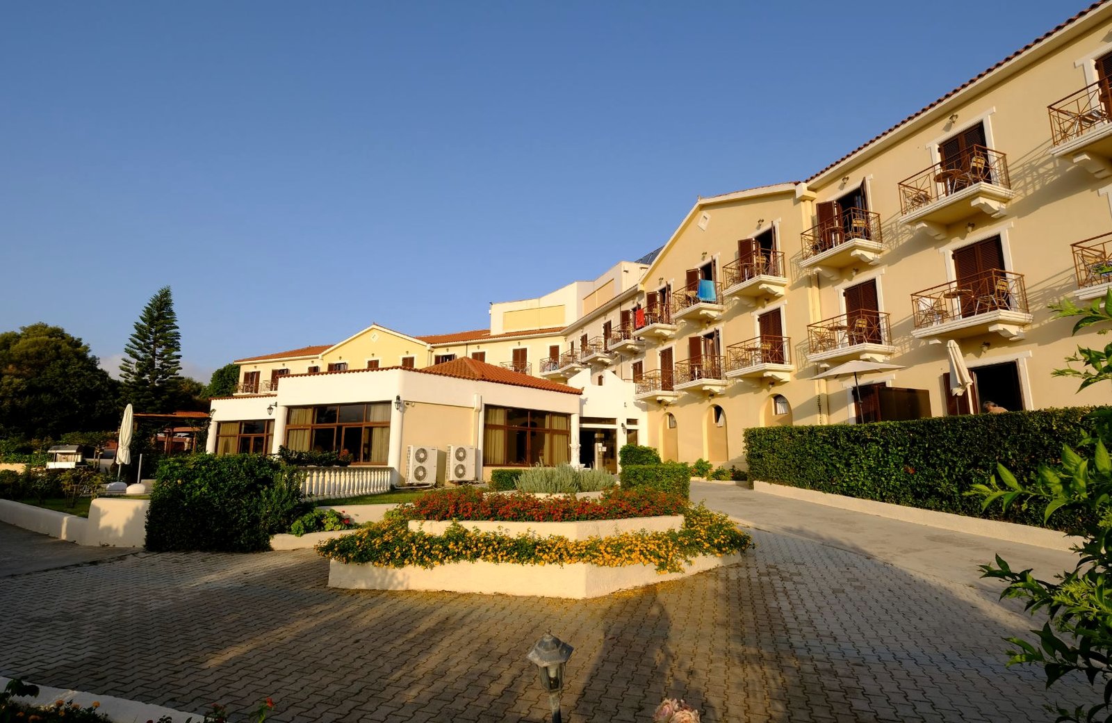 The cream low-rise buildings and covered restaurant terrace of the Karavados Beach Hotel above its entrance driveway and flower garden.