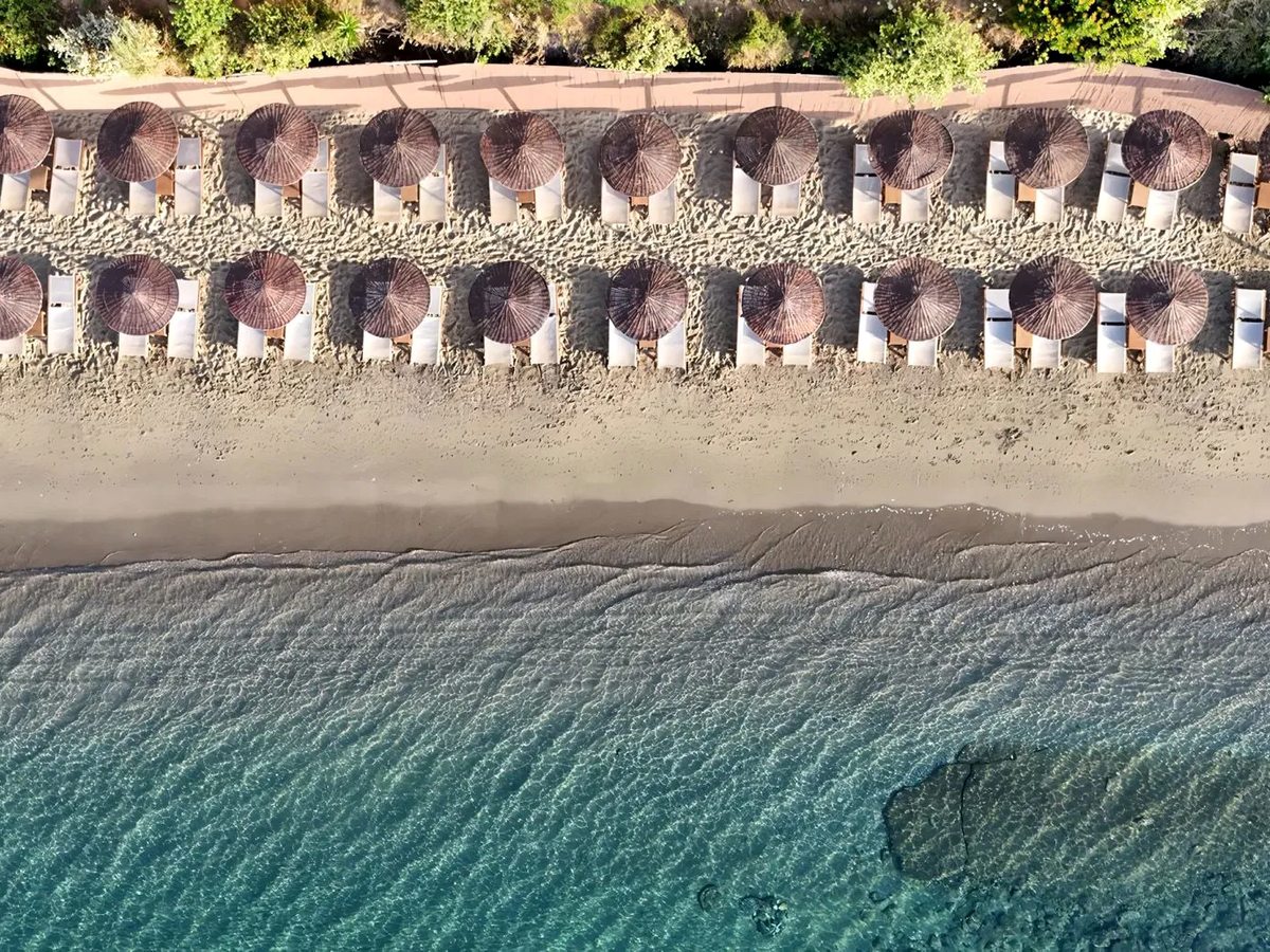 Aerial view of a sandy beach lined with rows of straw parasols and sun loungers beside turquoise shallows.