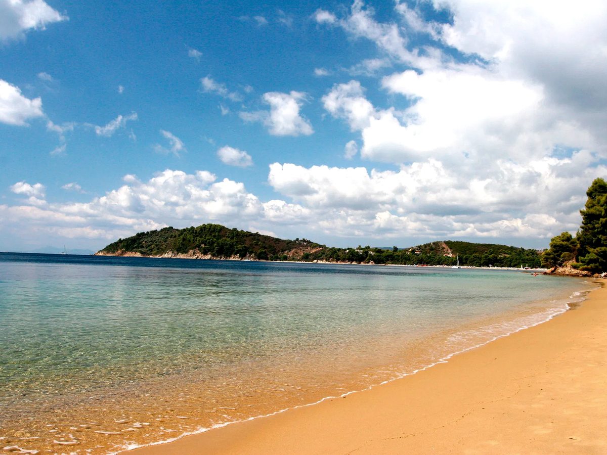 Sandy beach curving around a calm, shallow bay of clear turquoise water backed by green hills.