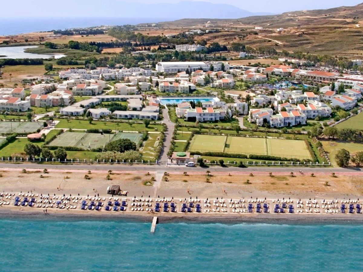 Aerial view of the resort's gardens, pool and sports pitches set back from a long pebble beach lined with blue sunbeds, separated by a coastal road beside a turquoise sea.