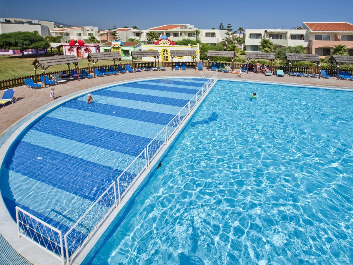A large curved pool with a fenced, blue-striped shallow section, blue loungers around it and colourful children's club buildings behind.