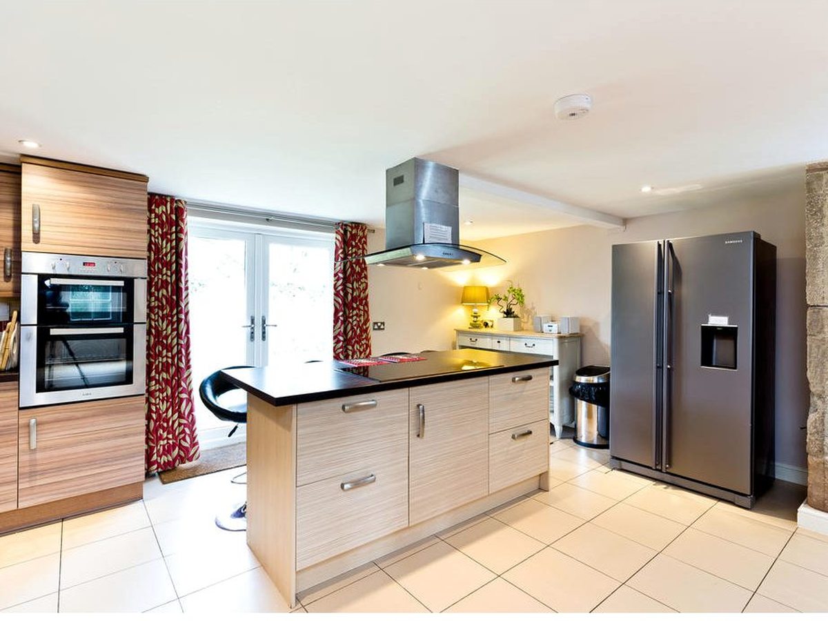 A cottage kitchen with pale wood units, a black-topped island with a hob and extractor above, a built-in double oven and a large American-style fridge freezer on a tiled floor.