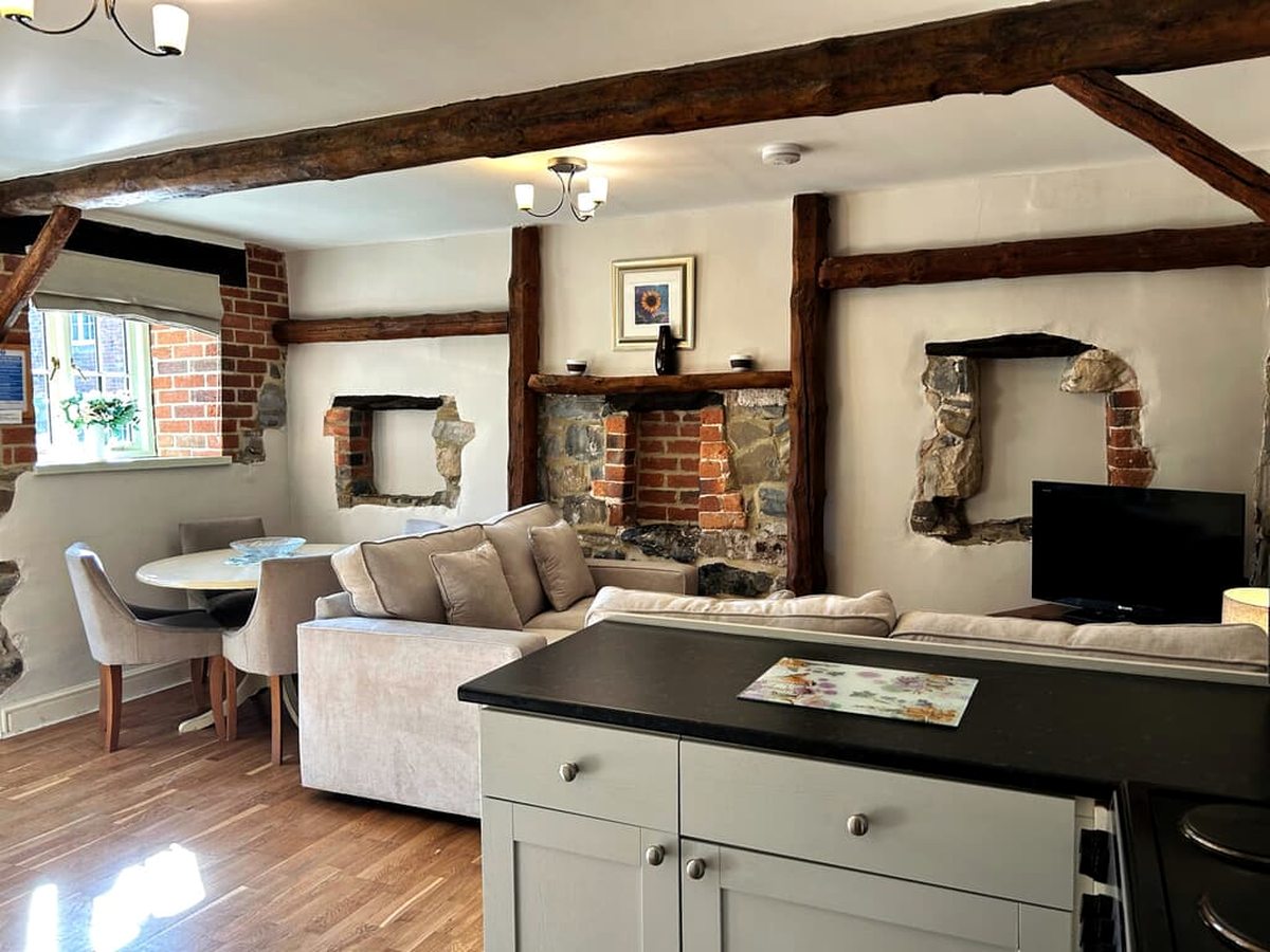 An open-plan cottage living area with heavy dark ceiling beams, exposed stone and brick openings in white walls, a cream corner sofa, a round dining table and a kitchen island in the foreground.