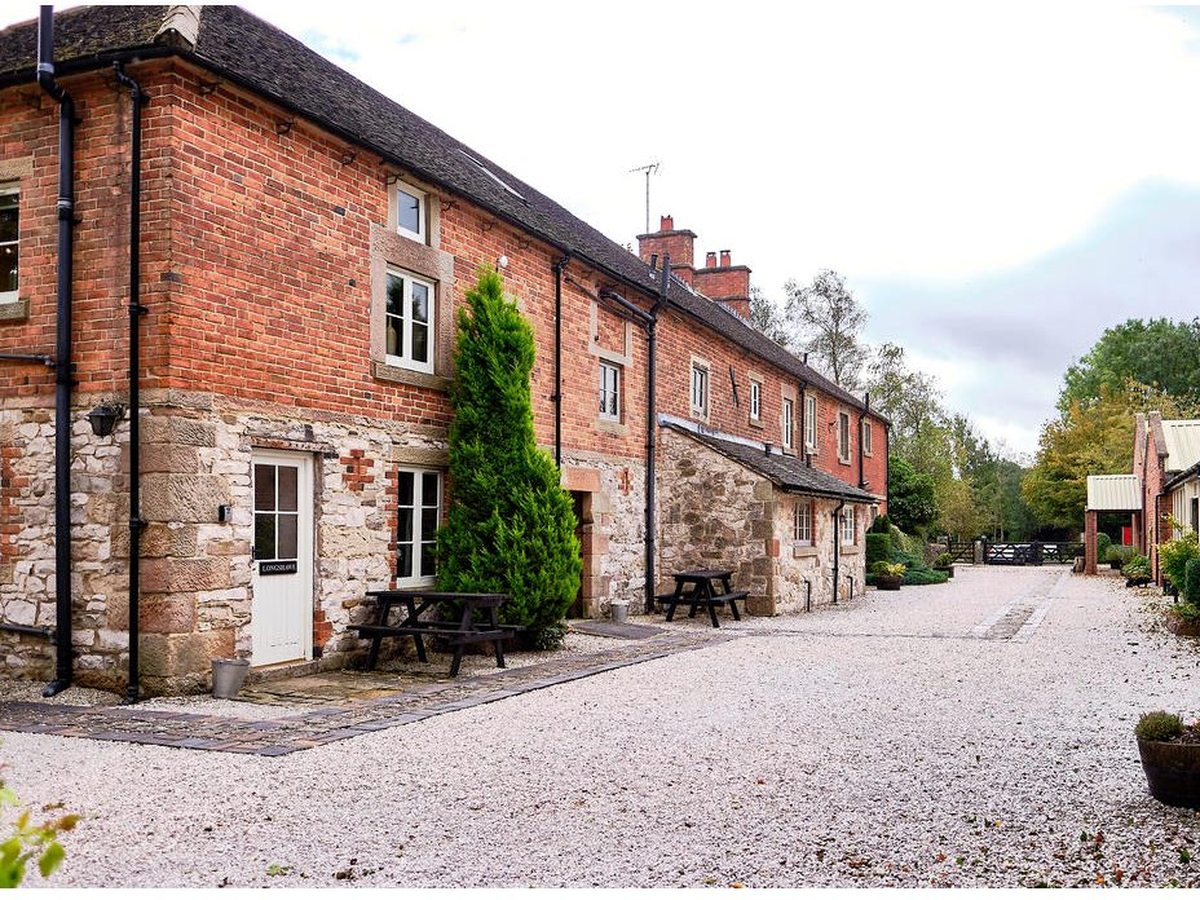 A terrace of brick and stone cottages along a gravel courtyard, with picnic benches and a white door outside each cottage and a clipped conifer against the wall.
