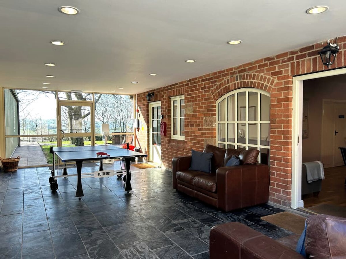 A games room with a blue table tennis table on a slate-tiled floor, brown leather sofas against an exposed brick wall and a glazed wall onto the grounds.