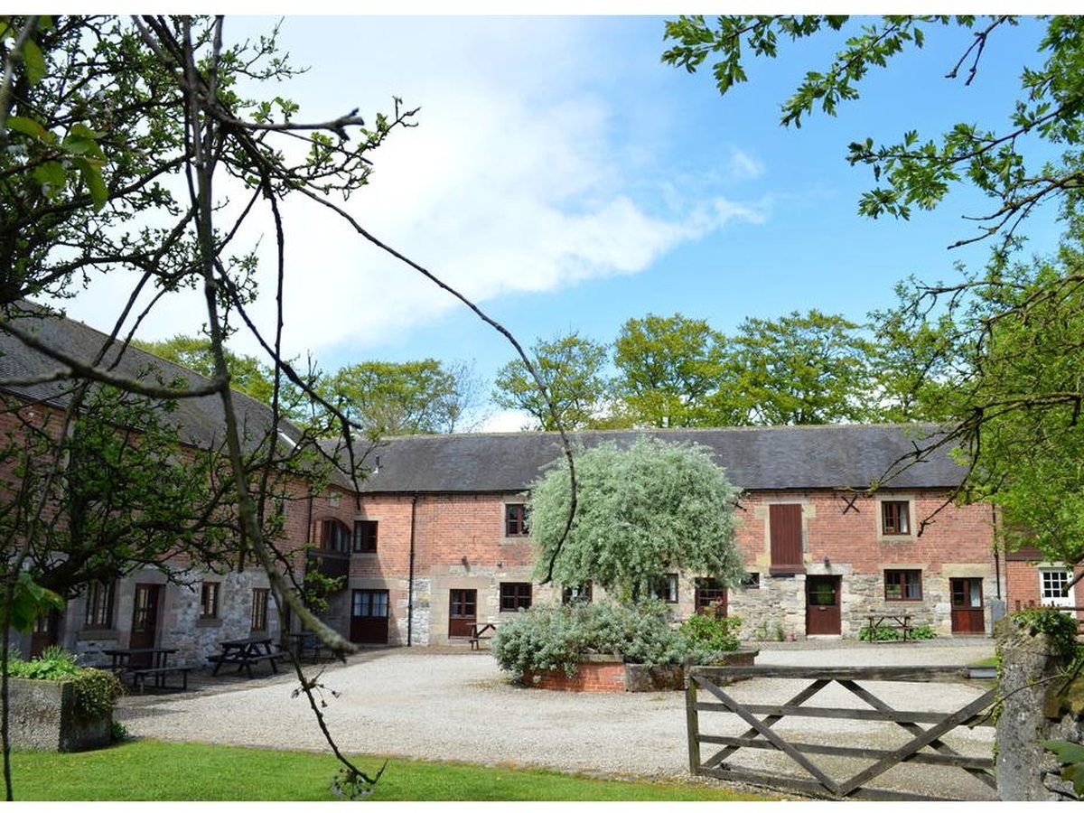 Two-storey brick and stone cottages ranged around a gravel courtyard with picnic benches, a clipped shrub in a raised bed and mature trees behind under a blue sky.