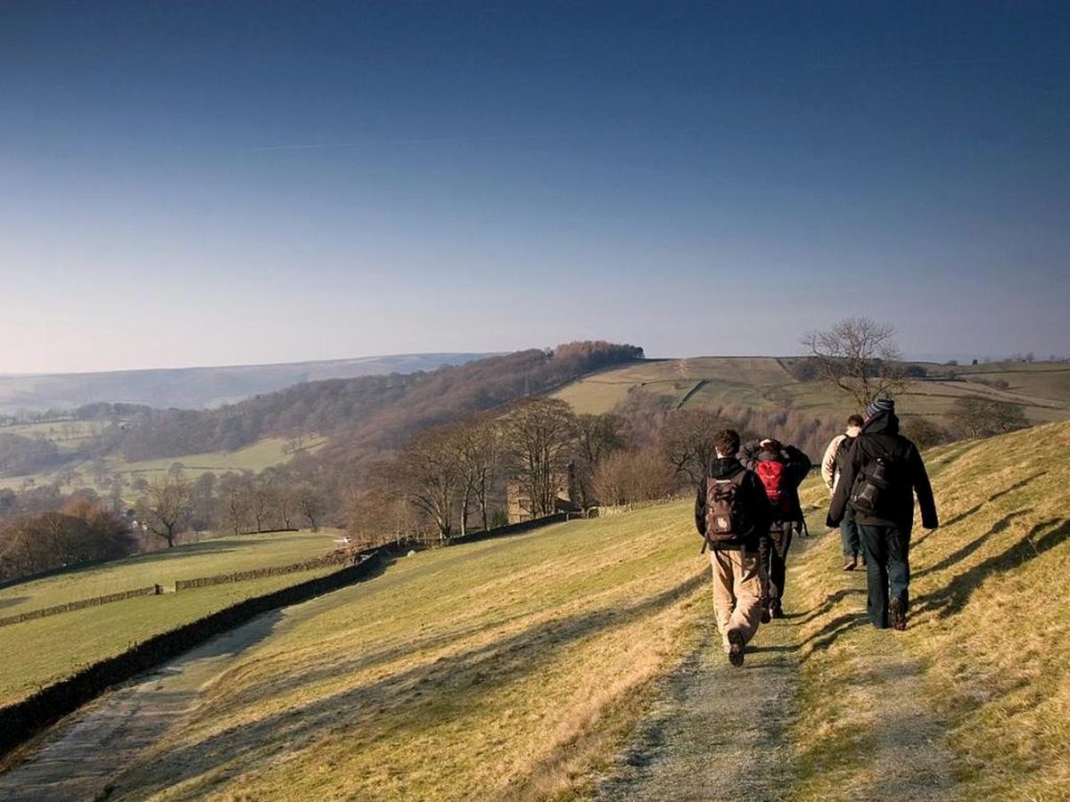 Four walkers with rucksacks on a grassy hillside path beside a drystone wall, looking across a wide valley of fields and bare winter trees under a clear blue sky.