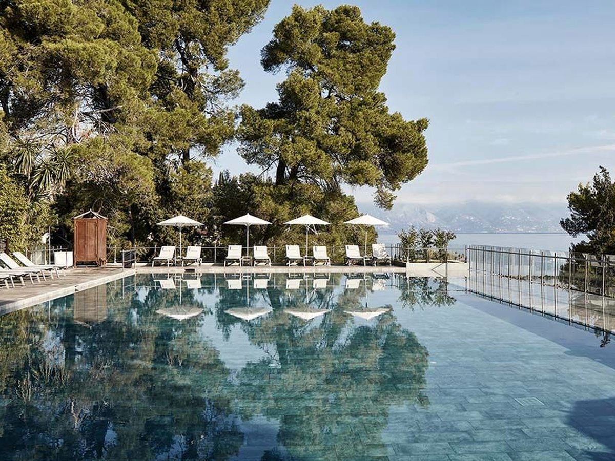 A long pool with a glass-railed infinity edge, a row of white parasols and loungers along one side under tall pines, and the sea and mountains beyond.