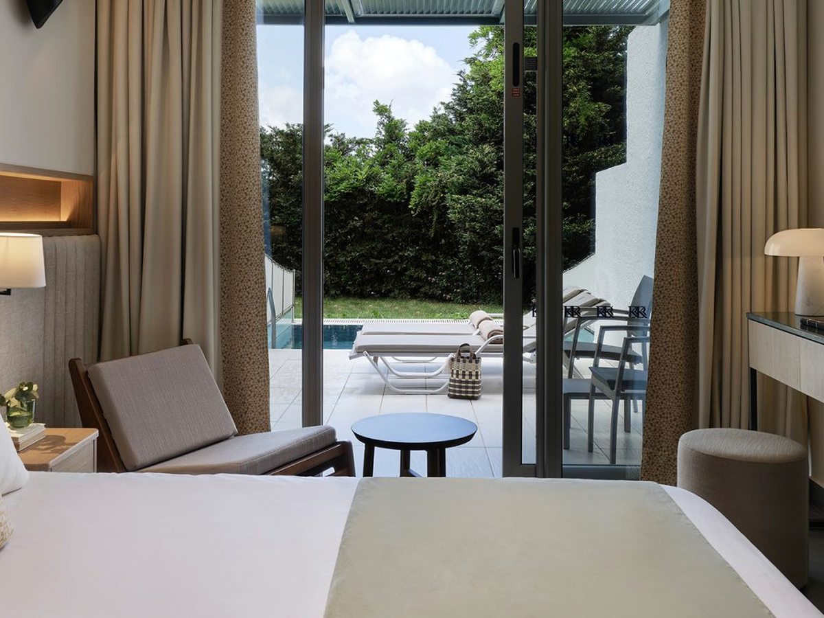 A bedroom with sliding doors open onto a paved terrace with a small blue pool, sun loungers, a table and chairs and a tall hedge behind.