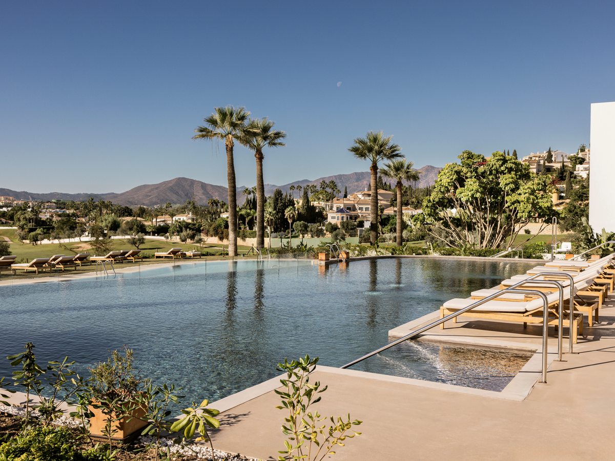 A large outdoor pool with an infinity edge, wooden sun loungers along one side, palm trees and mountains in the distance.