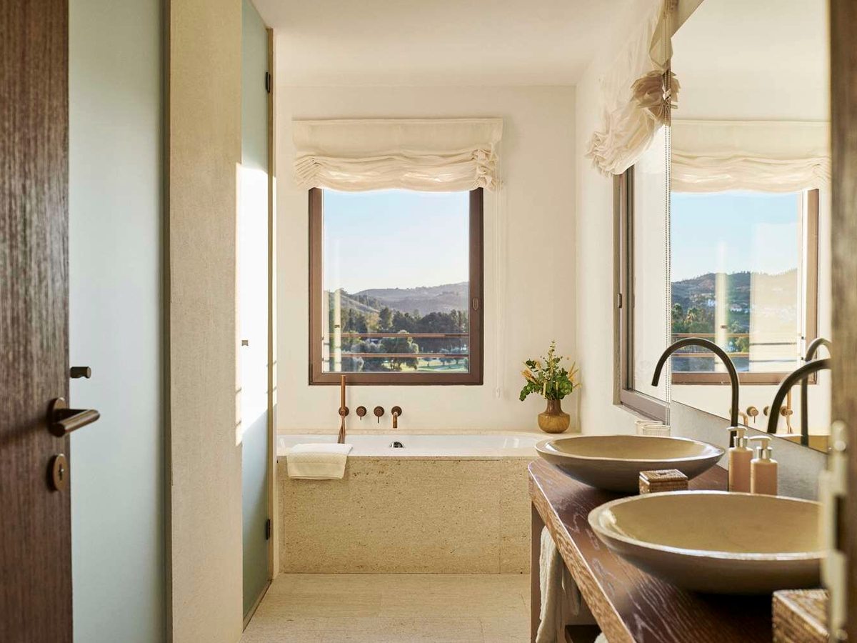 A bathroom with a stone-clad bath under a window facing hills, twin stone basins on a dark wood vanity and a large mirror.