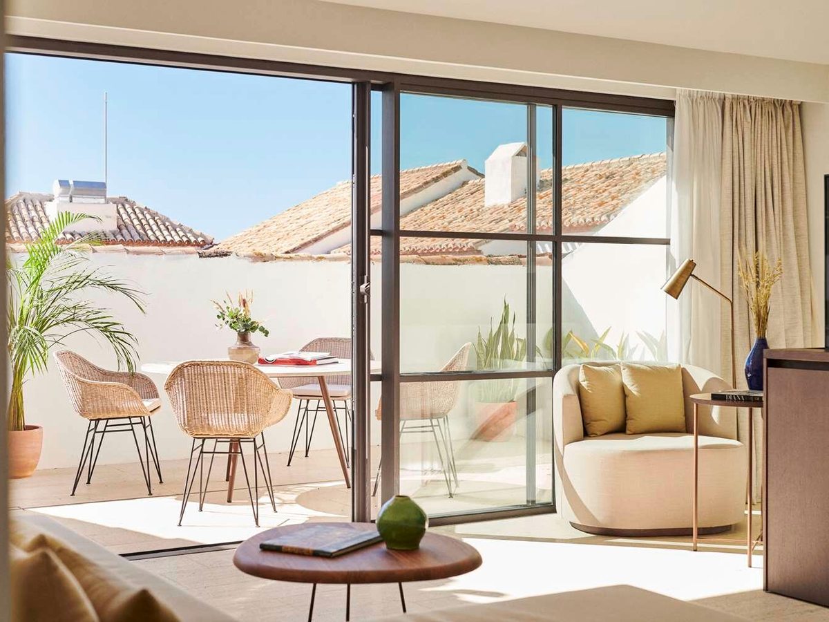 A suite living room opening through sliding doors onto a private terrace with a dining table, wicker chairs and potted palms, tiled roofs behind.