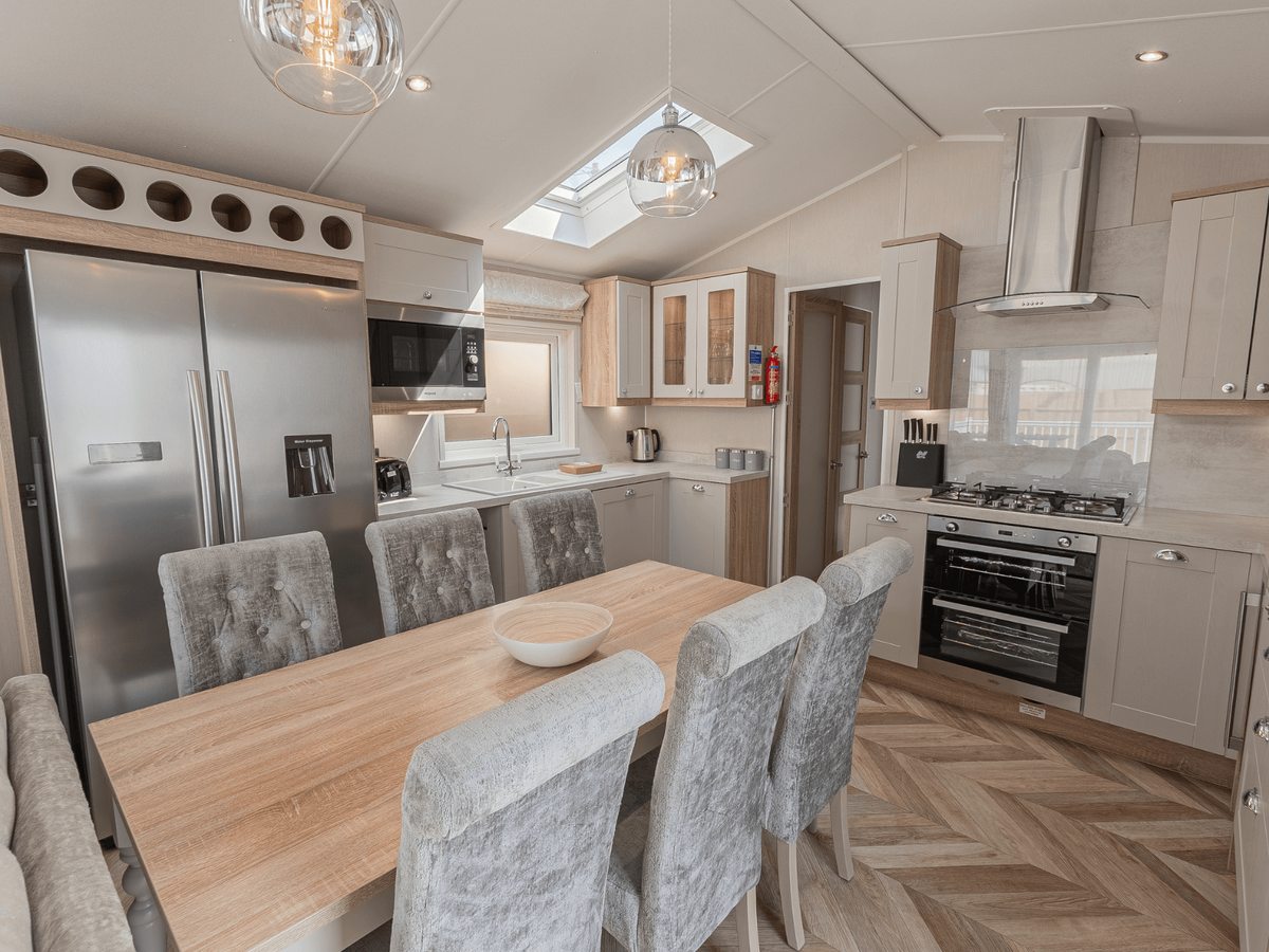 Open-plan kitchen with cream shaker units, a stainless American-style fridge freezer, a gas hob, oven, microwave and extractor, and a wooden dining table with six upholstered chairs under a skylight.