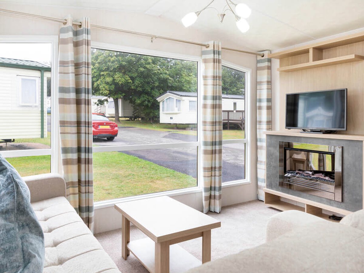 Caravan lounge with cream sofas along two walls, a pale wood coffee table, a wall-mounted TV above a gas fire and large windows onto a road and other caravans.