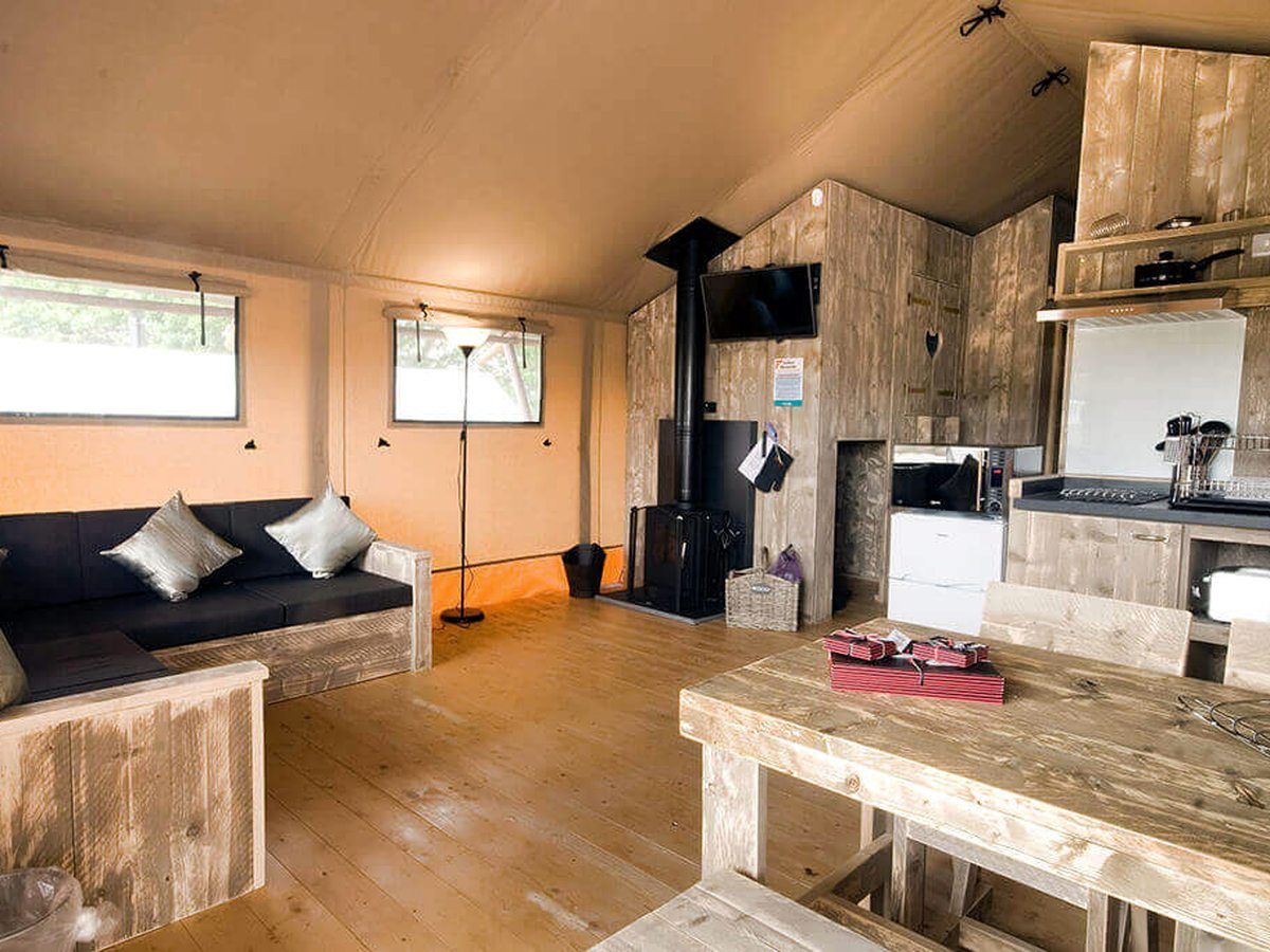 Canvas-walled safari tent interior with a timber-framed sofa, a wooden dining table and benches, a black log burner and a small kitchen with a fridge, microwave and hob along the back wall.