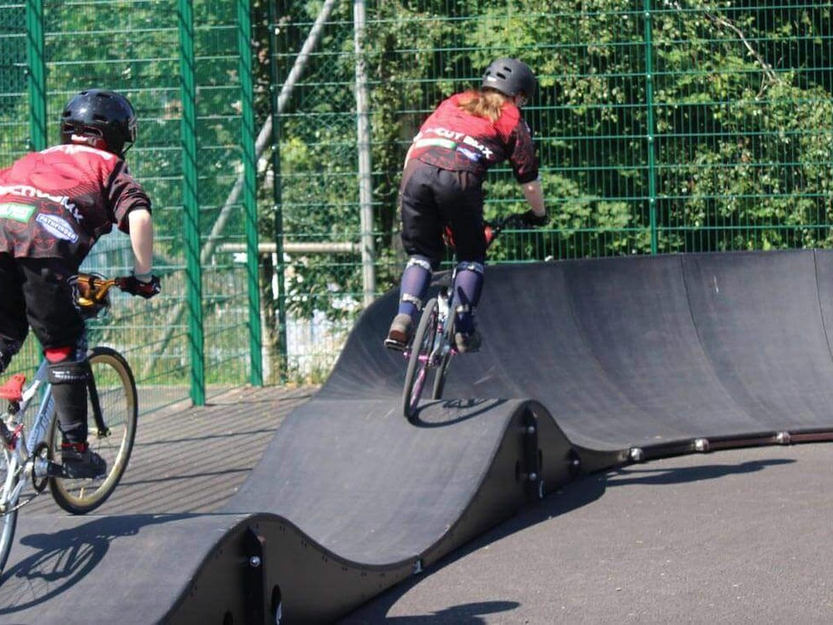 Two young riders in helmets and race kit riding over the rollers of a black moulded pump track inside a green mesh fence, with trees behind.