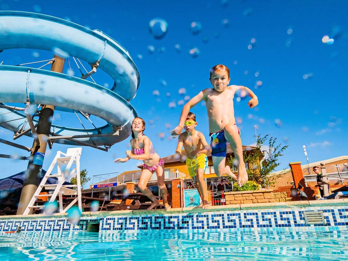 Three children jumping into the outdoor pool from the tiled poolside, with the blue spiral waterslide, a lifeguard chair and the reception building behind under a clear blue sky.