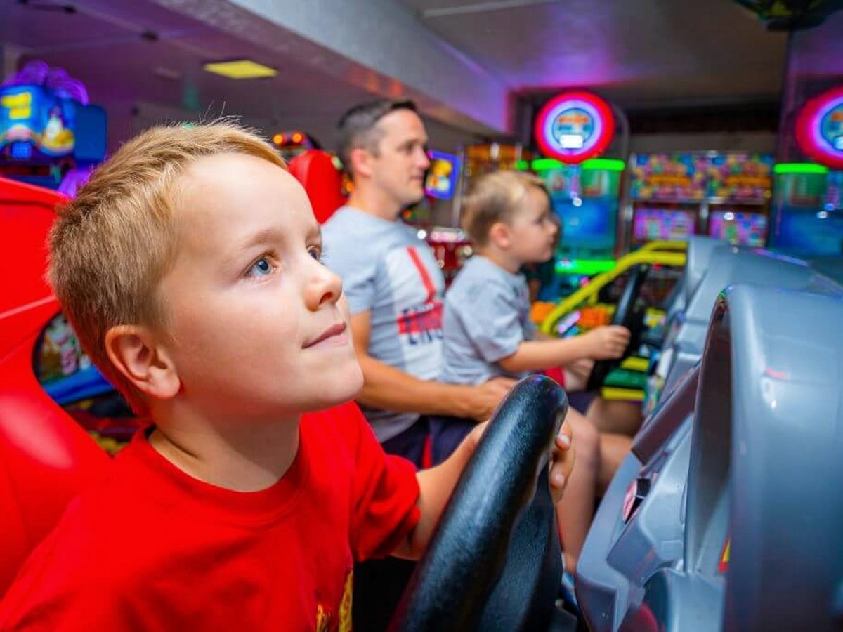A boy gripping the wheel of an arcade driving game while a man and a younger child play the machines beside him, in a dark games room lit by screens.