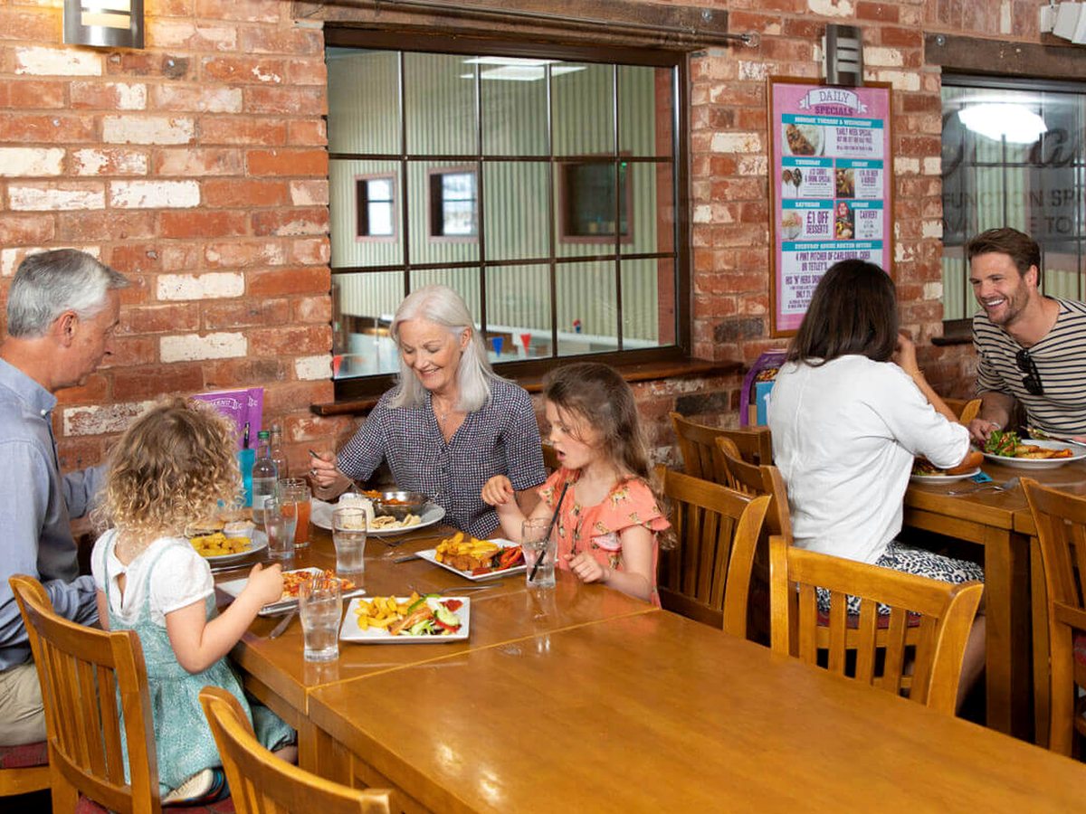 Two adults and two children eating plates of fish and chips at a long wooden table in a restaurant with exposed brick walls, wooden chairs and a specials poster on the wall.