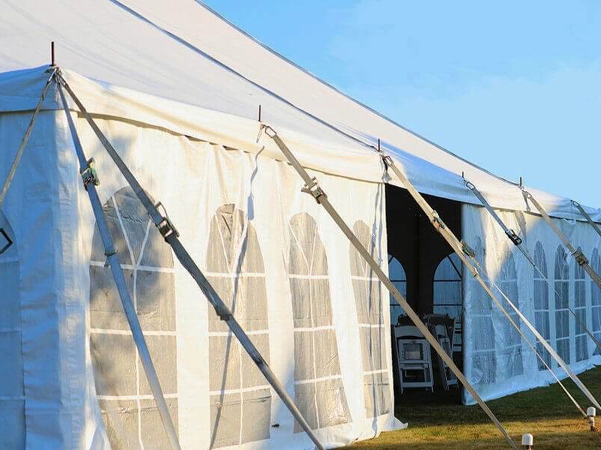 A large white marquee with arched window panels and guy ropes pitched on grass under a blue sky, with folded chairs stacked inside the open entrance.