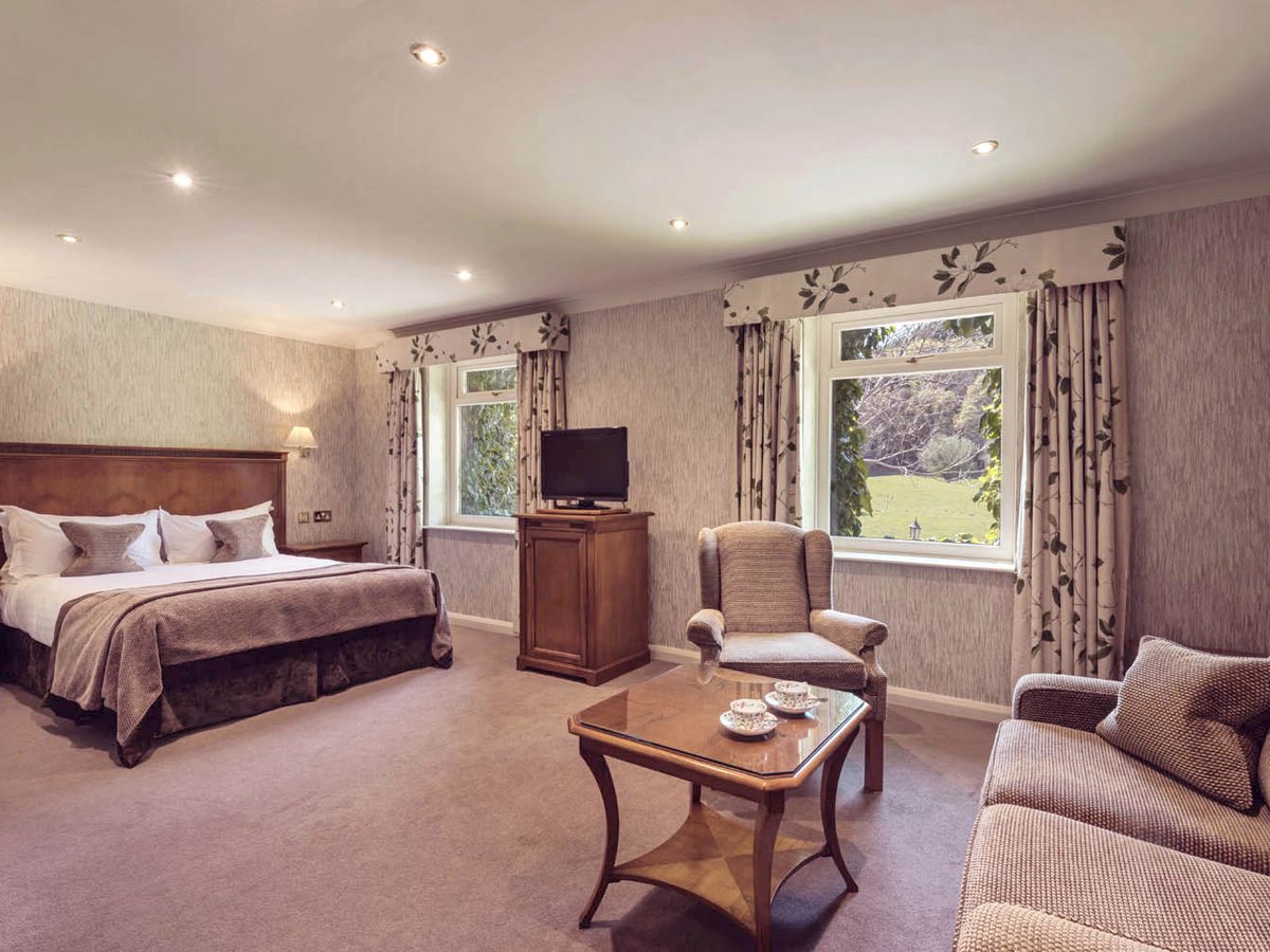 Bedroom with a wooden headboard, a sofa, a wing-back armchair, a coffee table and a television, with two curtained windows on to grass and trees.