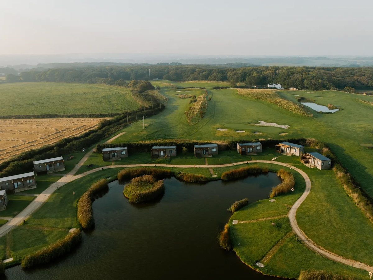 Aerial view of a reed-fringed lake with small lodges along one side, a green golf course with bunkers behind and farmland at the edges.