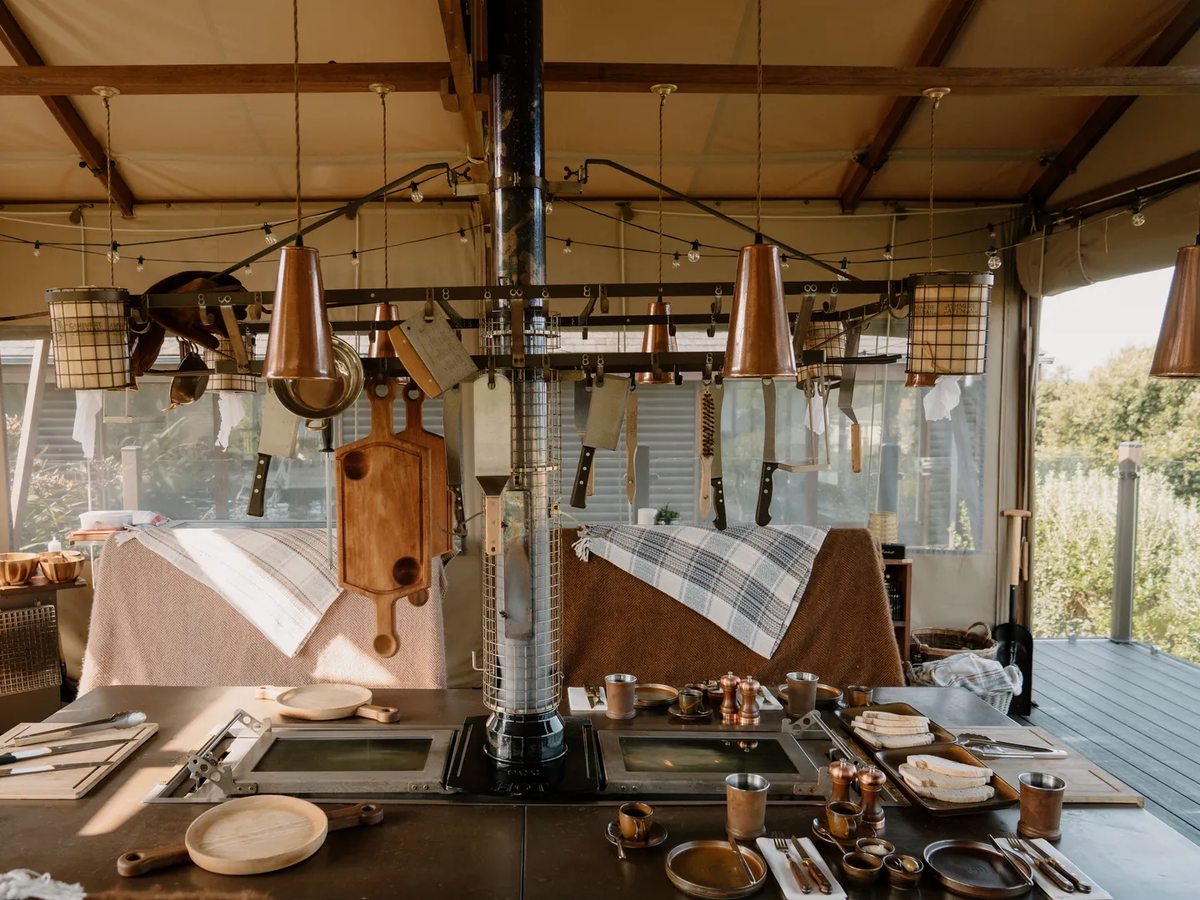 A long outdoor kitchen table with a central charcoal grill and flue, copper pendant lamps above and places set along both sides, under a canvas roof.