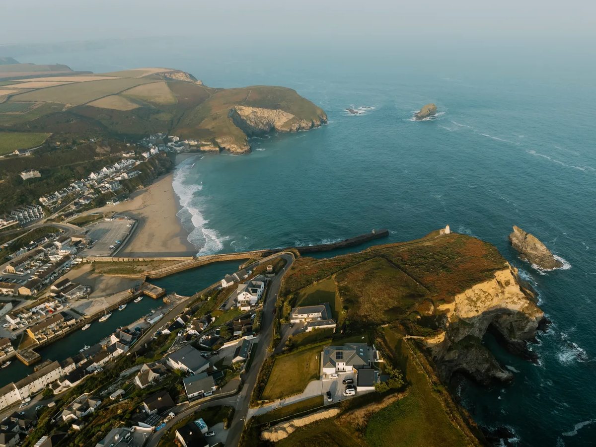 Aerial view of a coastal village at the mouth of a valley, with a stone harbour arm, a sandy beach and grassy headlands either side.