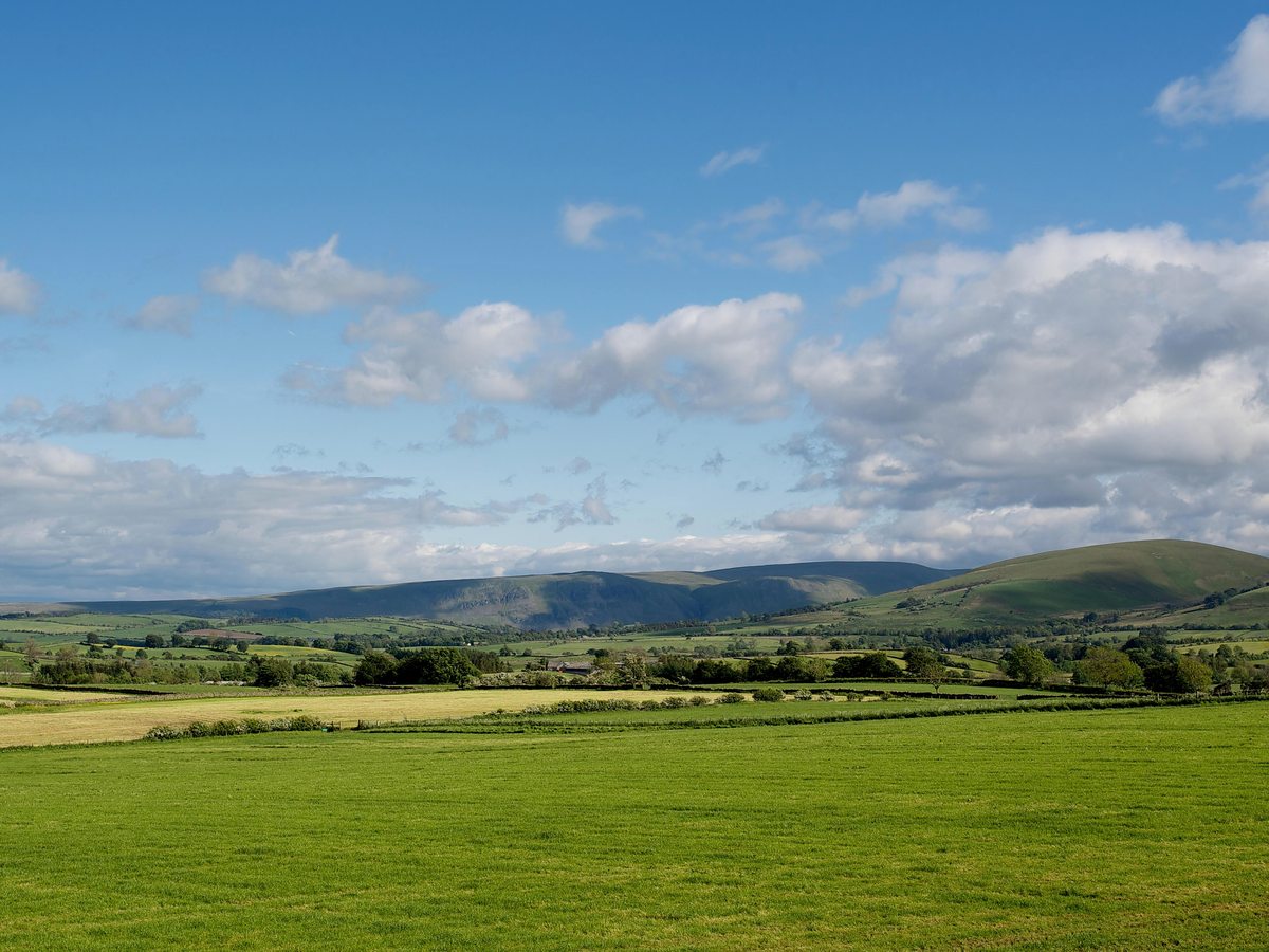 Green pasture and hedgerows in the foreground with a long line of low fells along the horizon under a blue sky with scattered cloud.