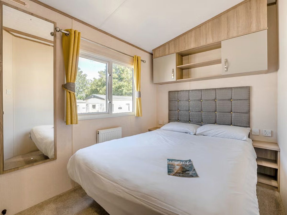 A double bedroom with a padded headboard, white bedding, overhead cupboards and a full-length mirror.