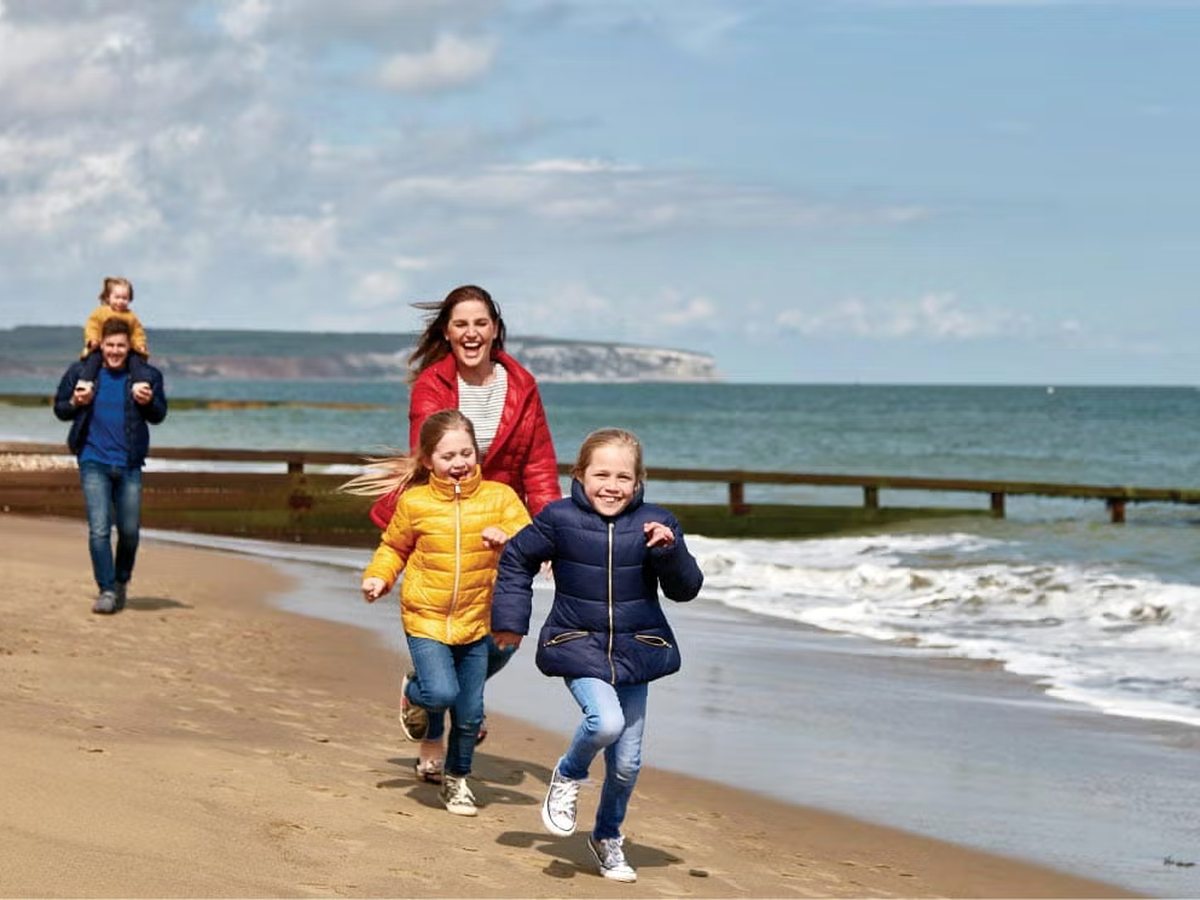Two children running along wet sand ahead of their parents, with breaking waves and a timber groyne behind.