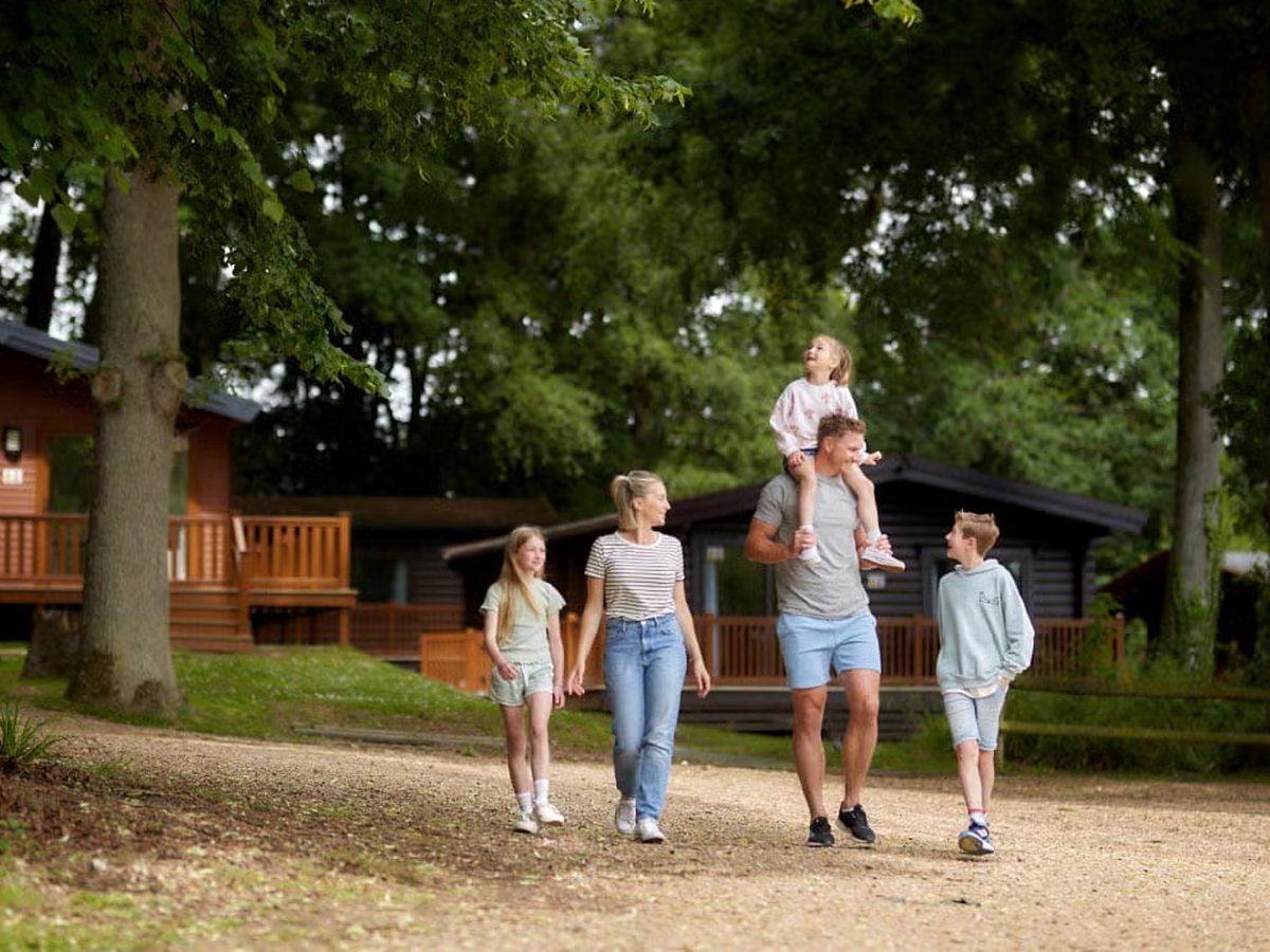A family walking along a gravel lane between timber lodges under mature trees.