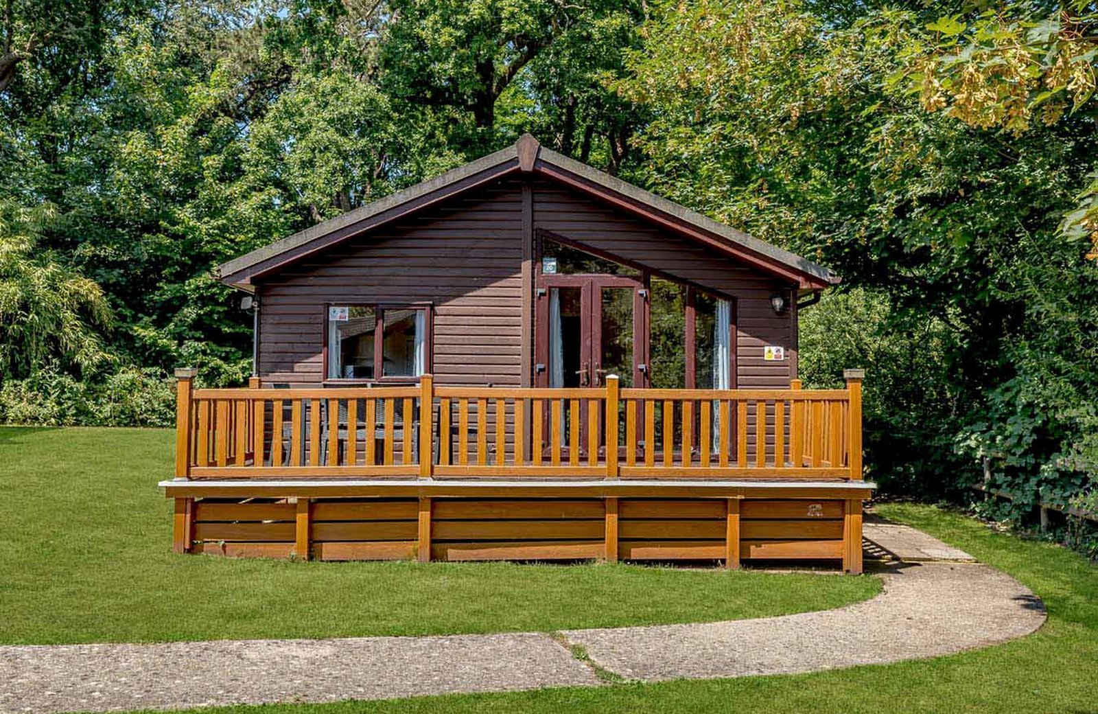 A timber lodge with a wooden veranda on a mown lawn, ringed by mature trees, at Landguard Holiday Park.