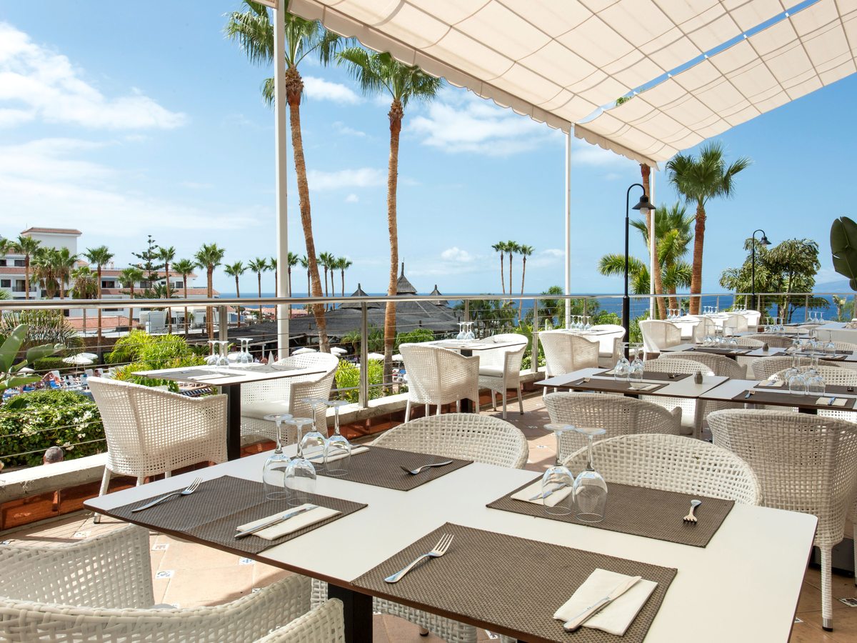 Covered outdoor restaurant terrace with white wicker chairs at set tables under a cream canopy, with palms and the sea beyond.
