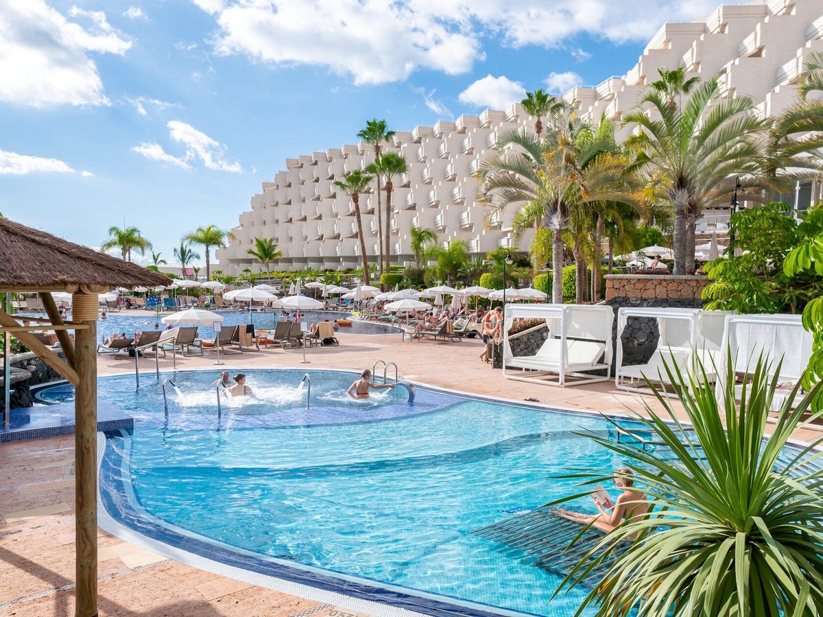 Curved shallow pool with hydromassage jets and guests sitting under them, with a larger pool, sun loungers and the terraced hotel building behind.