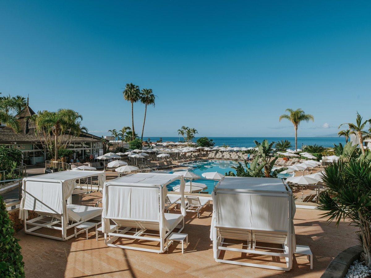 Three white curtained daybeds with canopies on a terracotta terrace above a curved pool ringed with parasols and loungers, with palms and the sea behind.