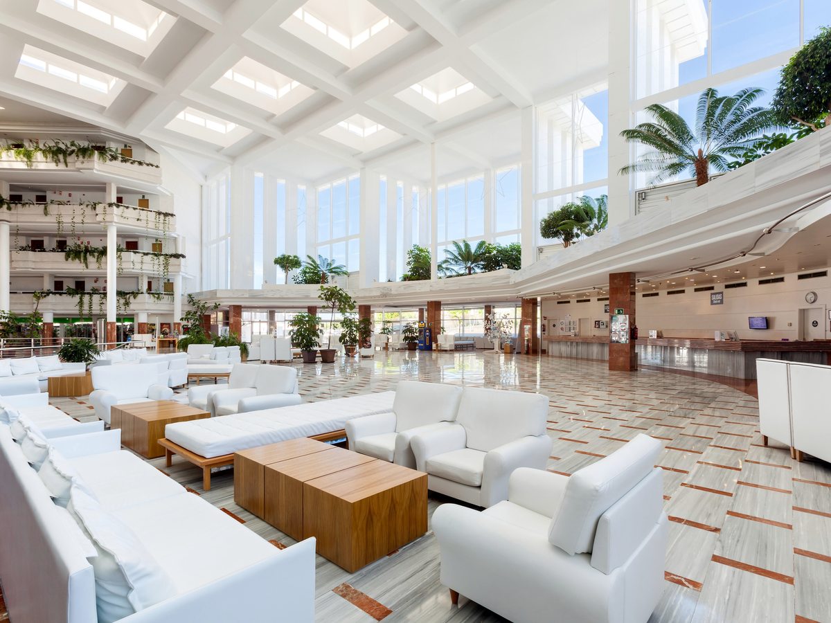 Bright open lobby with white sofas and armchairs around low wooden tables, a marble floor, tall windows and the reception desk along the far wall.