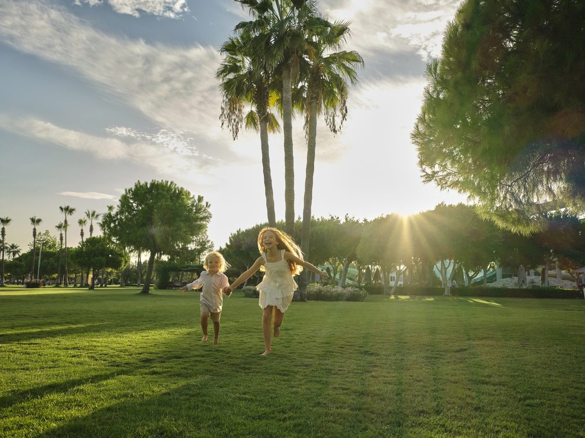 Two young children running across a wide green lawn towards tall palms, with low resort buildings behind and the sun low in a clear sky.